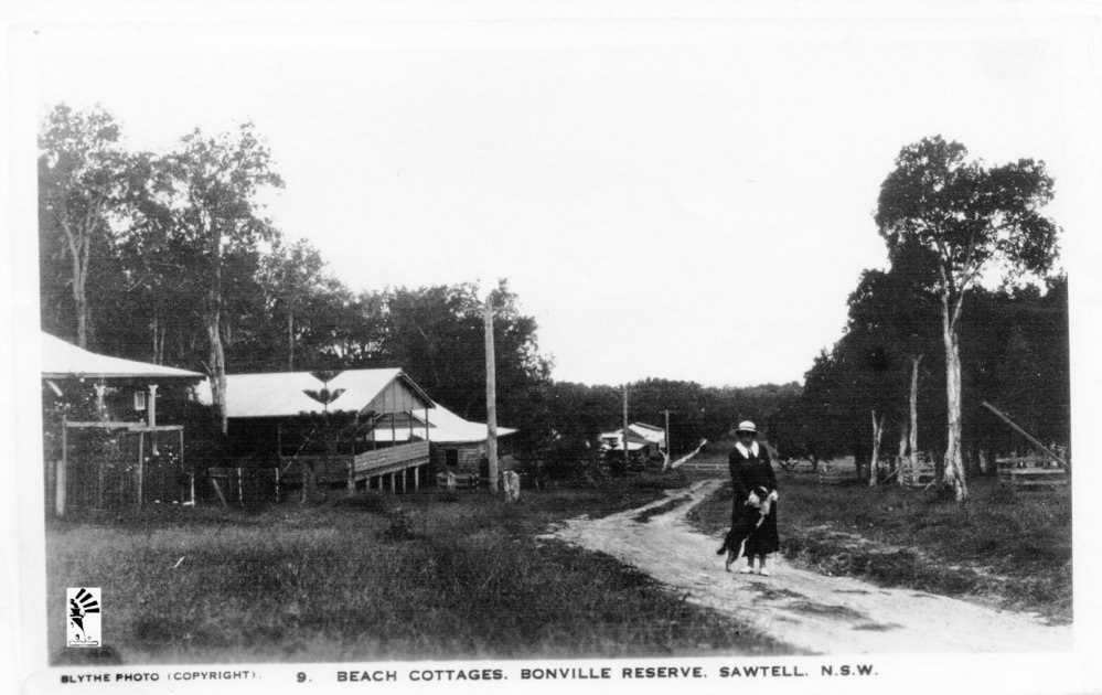Ethel Bailey walks her dog past the beach cottages at Bonville Reserve, 1930s
