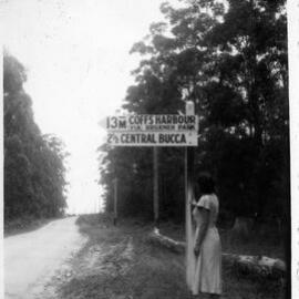 Ivy standing by the road sign 13 miles from Coffs Harbour, c.1955